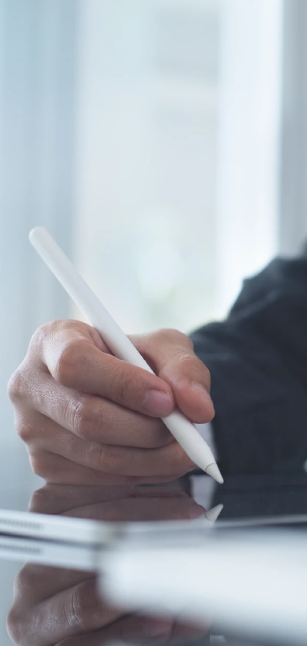 Eine Person hält einen Stylus und schreibt auf einem Tablet, während sie an einem Tisch sitzt. Im Hintergrund sind unscharfe Büromöbel sichtbar.