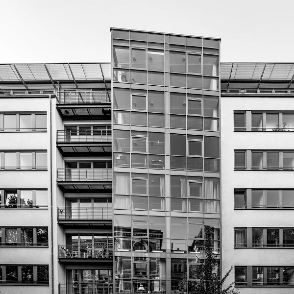 Ein modernes Wohngebäude mit einer markanten Glasfassade und mehreren Balkonen, das von einer hellen, neutralen Wand umgeben ist. Der obere Bereich des Gebäudes hat eine schräge Dachkonstruktion.