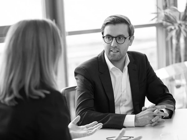 Ein Mann im Anzug sitzt an einem Tisch und hört aufmerksam einer Frau zu, die mit gestikulierenden Händen spricht. Im Hintergrund sind große Fenster und eine grüne Pflanze sichtbar.