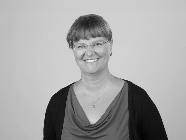 Eine lächelnde Frau mit kurzen, grauen Haaren und Brille trägt ein rotes Oberteil und einen schwarzen Cardigan. Sie steht vor einem neutralen, hellgrauen Hintergrund.