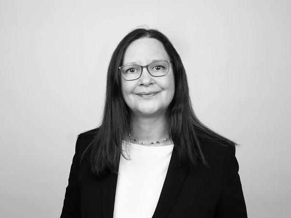 Eine Person mit langen, glatten, braunen Haaren und Brille lächelt freundlich in die Kamera, trägt einen schwarzen Blazer und ein weißes Oberteil vor einem neutralen Hintergrund.