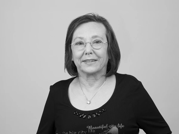 Eine lächelnde ältere Frau mit kurzen, grauen Haaren trägt eine schwarze Bluse mit Schriftzug und Schmuck. Der Hintergrund ist neutral grau.