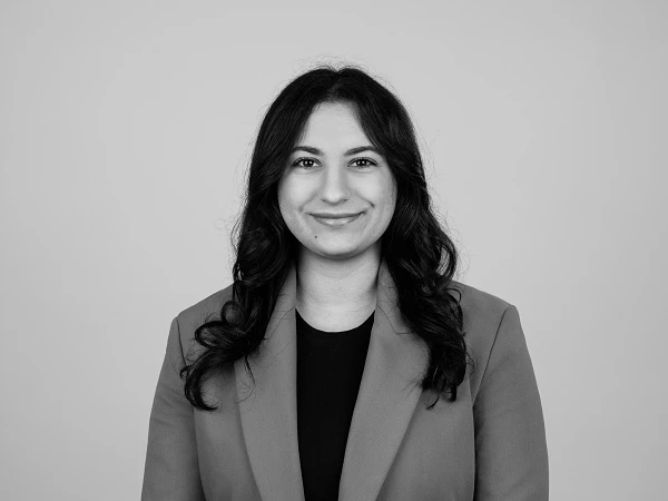 Eine lächelnde Frau mit langen, lockigen Haaren steht vor einem einfarbigen Hintergrund und trägt einen Blazer.