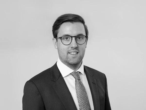 Ein gut gekleideter Mann mit Brille trägt einen Anzug und eine Krawatte mit geometrischem Muster. Er lächelt freundlich in die Kamera, vor einem neutralen, hellen Hintergrund.