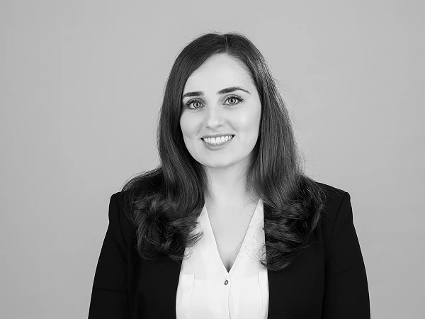 Eine lächelnde Frau mit langen, lockigen Haaren trägt ein schwarzes Blazer und ein weißes Hemd vor einem neutralen, hellen Hintergrund.