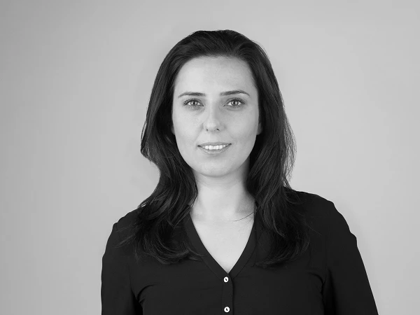 Eine Frau mit langen, dunklen Haaren und einem freundlichen Lächeln trägt ein schwarzes Oberteil. Der Hintergrund ist neutral und grau.