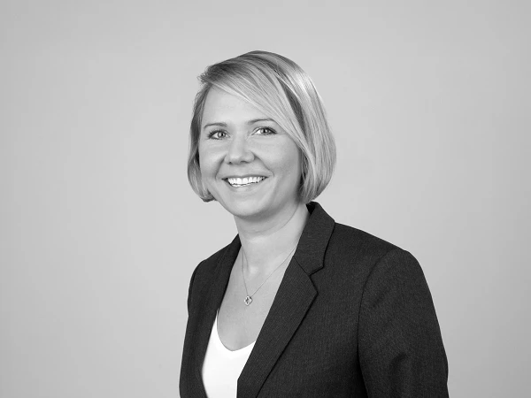 Eine lächelnde Frau mit blonden, schulterlangen Haaren trägt ein schwarzes Blazer-Jackett und eine weiße Bluse. Der Hintergrund ist grau.