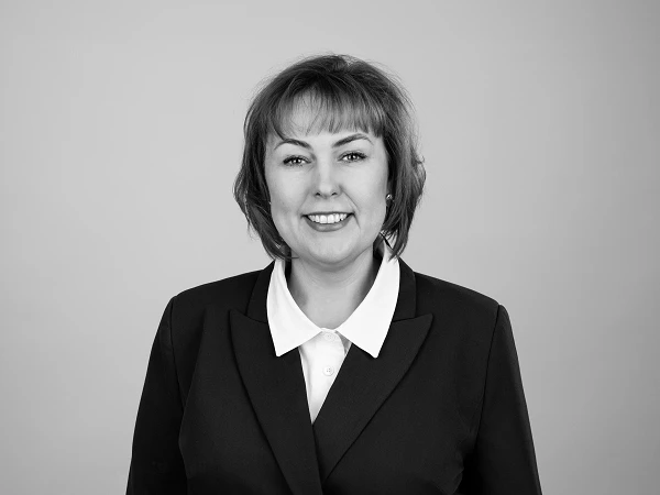 Eine lächelnde Frau mit schulterlangen, braunen Haaren trägt einen schwarzen Blazer und ein weißes Hemd. Der Hintergrund ist einfarbig und hell.