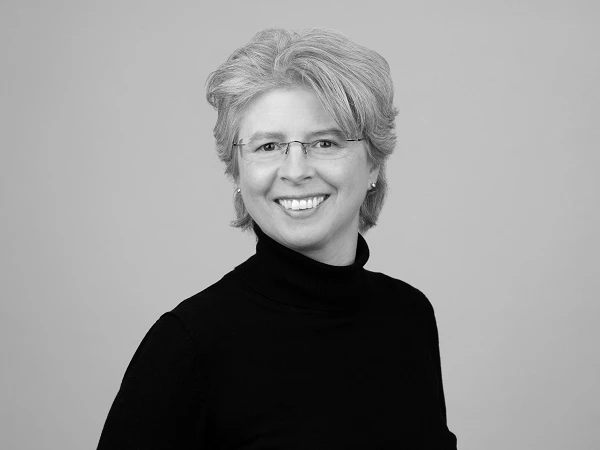 Eine lächelnde Frau mit kurzem, blonden Haar und Brille trägt einen schwarzen Rollkragenpullover und steht vor einem grauen Hintergrund.