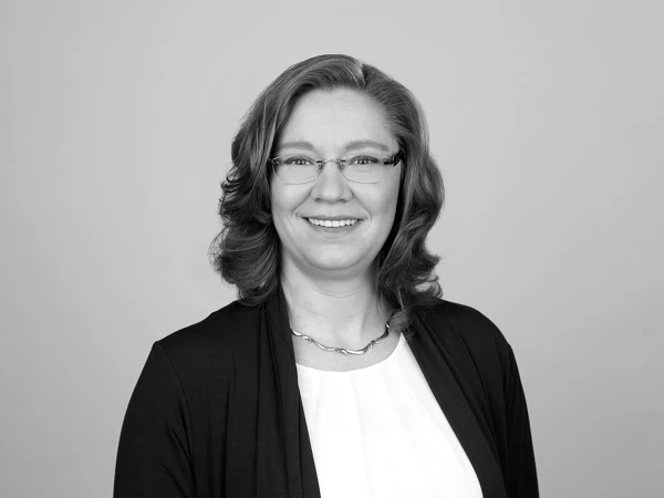 Eine freundlich lächelnde Frau mit langen, lockigen Haaren trägt eine weiße Bluse und einen dunklen Cardigan. Sie steht vor einem neutralen, hellgrauen Hintergrund.