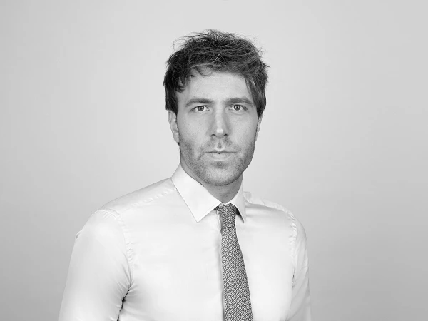 Ein junger Mann in einem hellen Hemd mit einem gestreiften roten Krawatte steht vor einem grauen Hintergrund und schaut direkt in die Kamera.