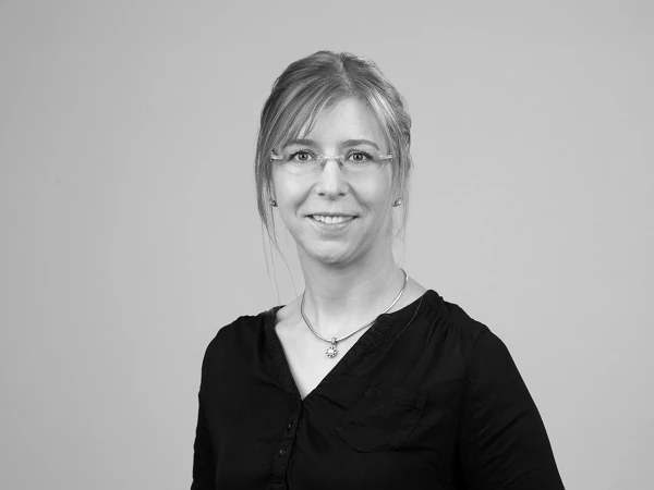 Eine Person mit langen, blonden Haaren und Brille trägt ein schwarzes Oberteil und lächelt freundlich in die Kamera vor einem grauen Hintergrund.