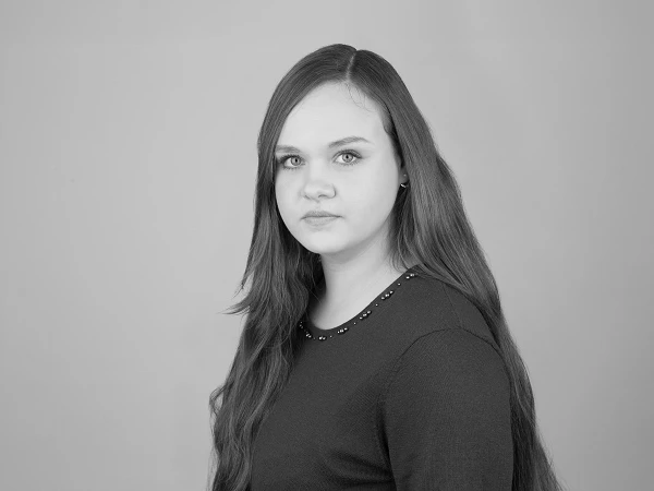 Eine junge Frau mit langen, glatten Haaren und einem neutralen Gesichtsausdruck schaut zur Seite. Sie trägt ein burgunderfarbiges Oberteil und steht vor einem hellgrauen Hintergrund.