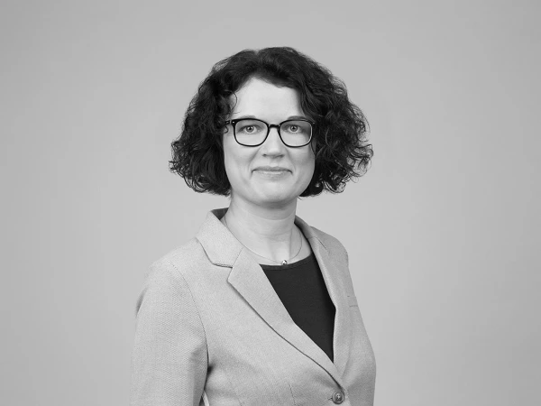 Eine Person mit lockigem, braunen Haar und Brille trägt einen grauen Blazer und blickt freundlich in die Kamera. Der Hintergrund ist neutral und einfarbig.