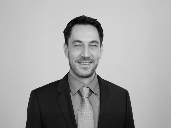 Ein Mann mit dunklen Haaren trägt einen schwarzen Anzug und ein hellblaues Hemd mit einer hellgrauen Krawatte und lächelt freundlich vor einem grauen Hintergrund.