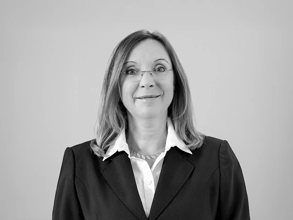 Eine Frau mit langen, blonden Haaren und Brille trägt einen schwarzen Blazer über einem weißen Hemd. Sie lächelt und steht vor einem grauen Hintergrund.