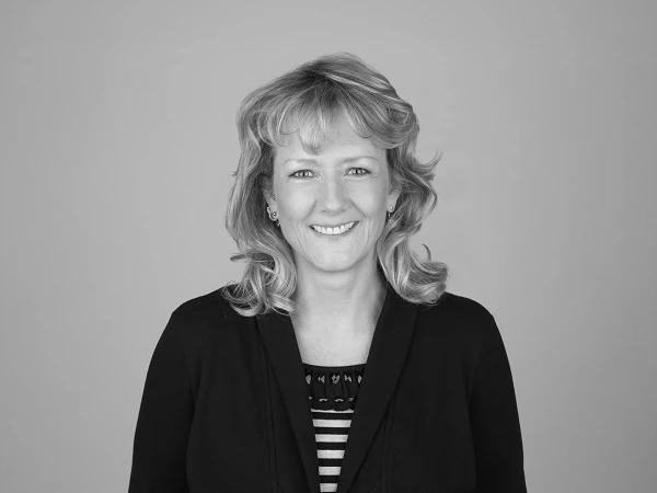 Eine lächelnde Frau mit blonden, leicht gewellten Haaren trägt einen schwarzen Pullover über einem gestreiften Oberteil und steht vor einem grauen Hintergrund.