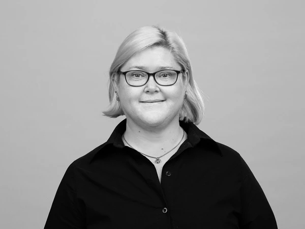 Eine lächelnde Person mit blonden, schulterlangen Haaren und einer schwarzen Bluse steht vor einem grauen Hintergrund. Sie trägt eine Brille und hat eine entspannte Ausstrahlung.
