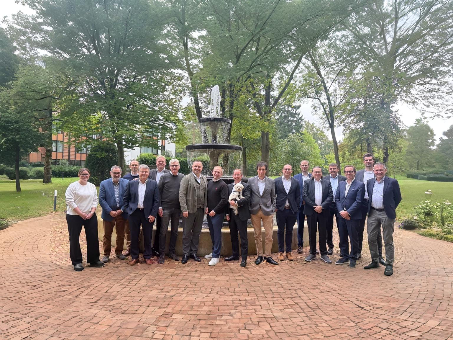 Eine Gruppe von 22 Personen steht um einen Brunnen in einem parkähnlichen Umfeld, umgeben von Bäumen und Grünflächen. Die Menschen lächeln und posieren für ein Gruppenfoto.