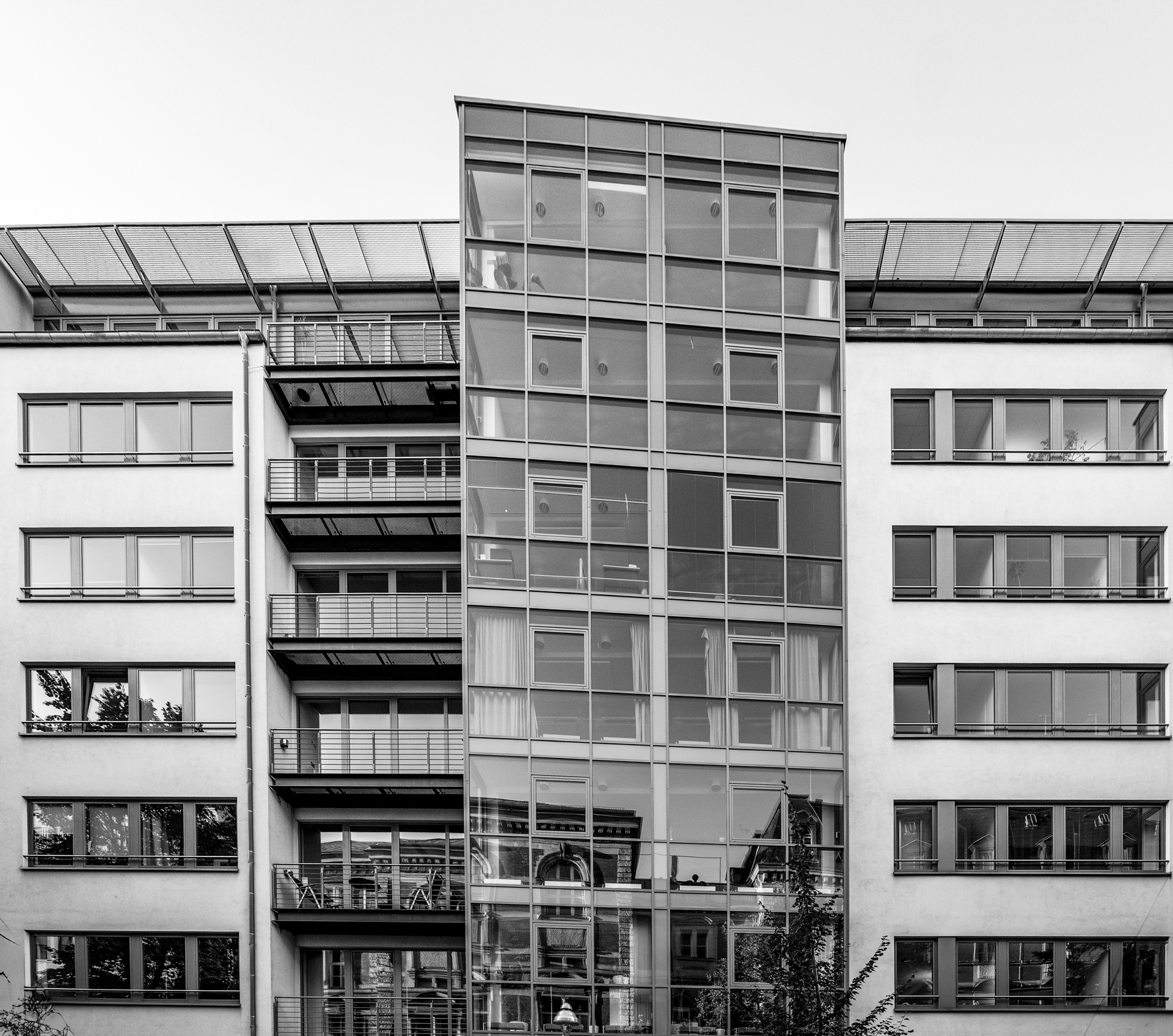 Ein modernes Wohngebäude mit einer markanten Glasfassade und mehreren Balkonen, das von einer hellen, neutralen Wand umgeben ist. Der obere Bereich des Gebäudes hat eine schräge Dachkonstruktion.