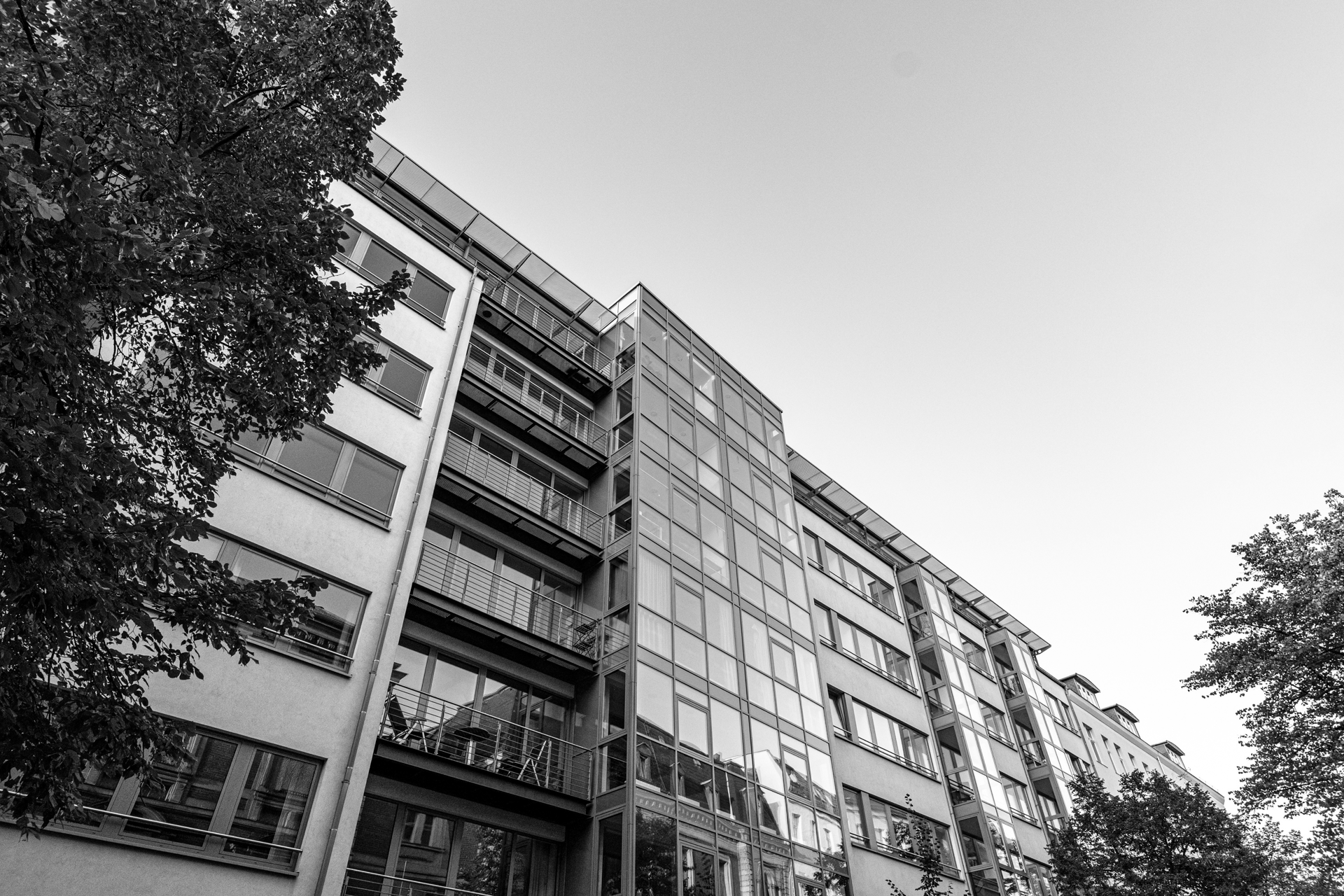 Moderne Wohngebäude mit großen Glasfenstern und Balkonen, umgeben von Bäumen, vor einem klaren blauen Himmel.