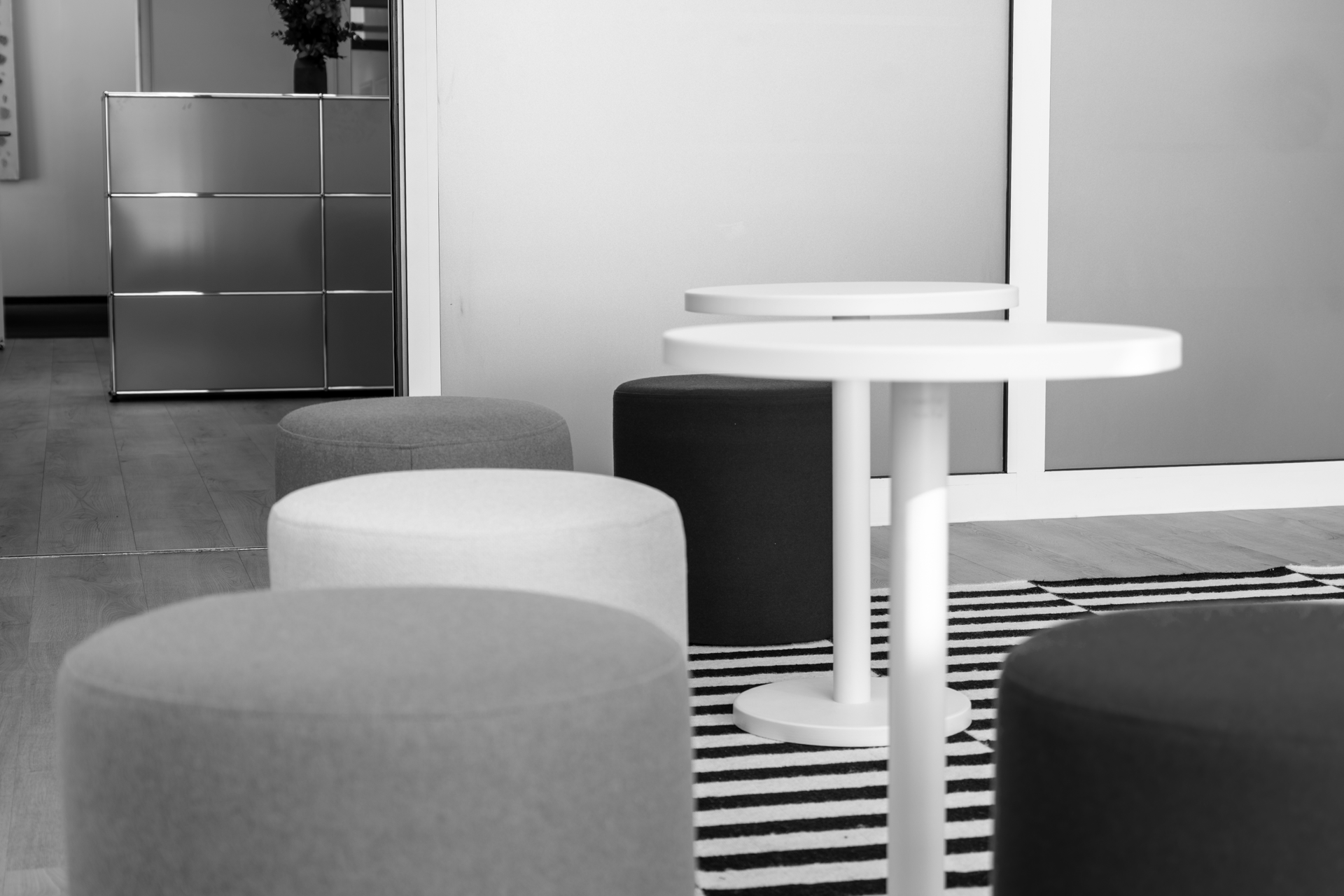 Bunte Sitzwürfel in Orange, Grau und Blau um einen weißen Tisch auf einem gestreiften Teppich in einem modernen Innenraum. Im Hintergrund ist eine silberne Kommode zu sehen.