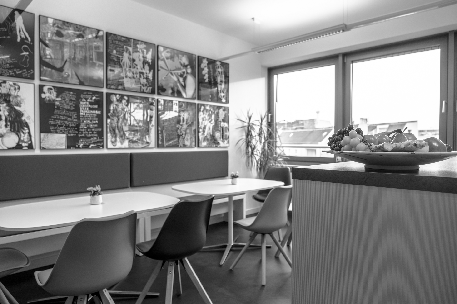 Ein moderner Aufenthaltsraum mit mehreren Tischen und Stühlen in verschiedenen Farben, einer Obstschale mit Äpfeln, Trauben und anderen Früchten auf einem Tisch, sowie einer Wand mit modernen Kunstwerken.