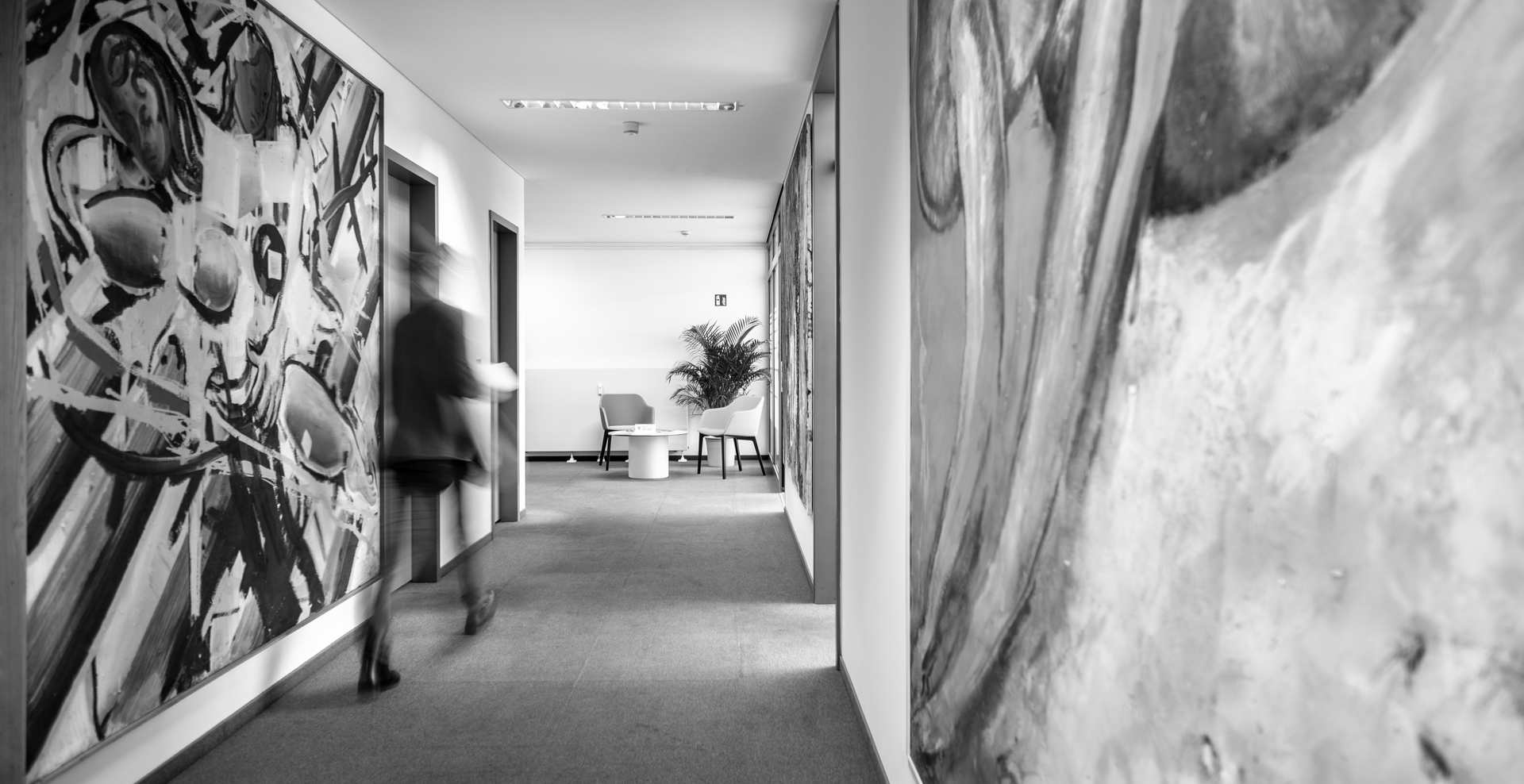 Ein Flur mit bunten, modernen Wandmalereien führt zu einem hellen Sitzbereich, in dem ein paar Möbelstücke stehen. Eine Person geht schnell durch den Flur.