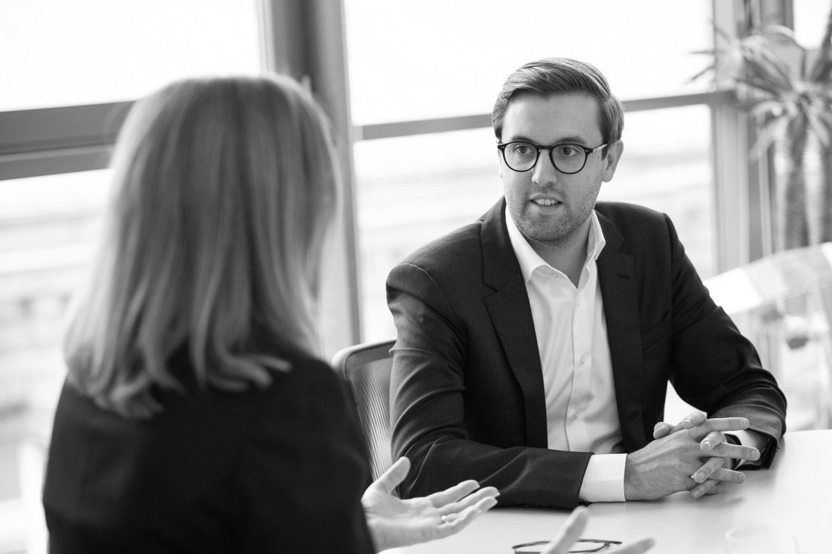 Ein Mann im Anzug sitzt an einem Tisch und hört aufmerksam einer Frau zu, die mit gestikulierenden Händen spricht. Im Hintergrund sind große Fenster und eine grüne Pflanze sichtbar.