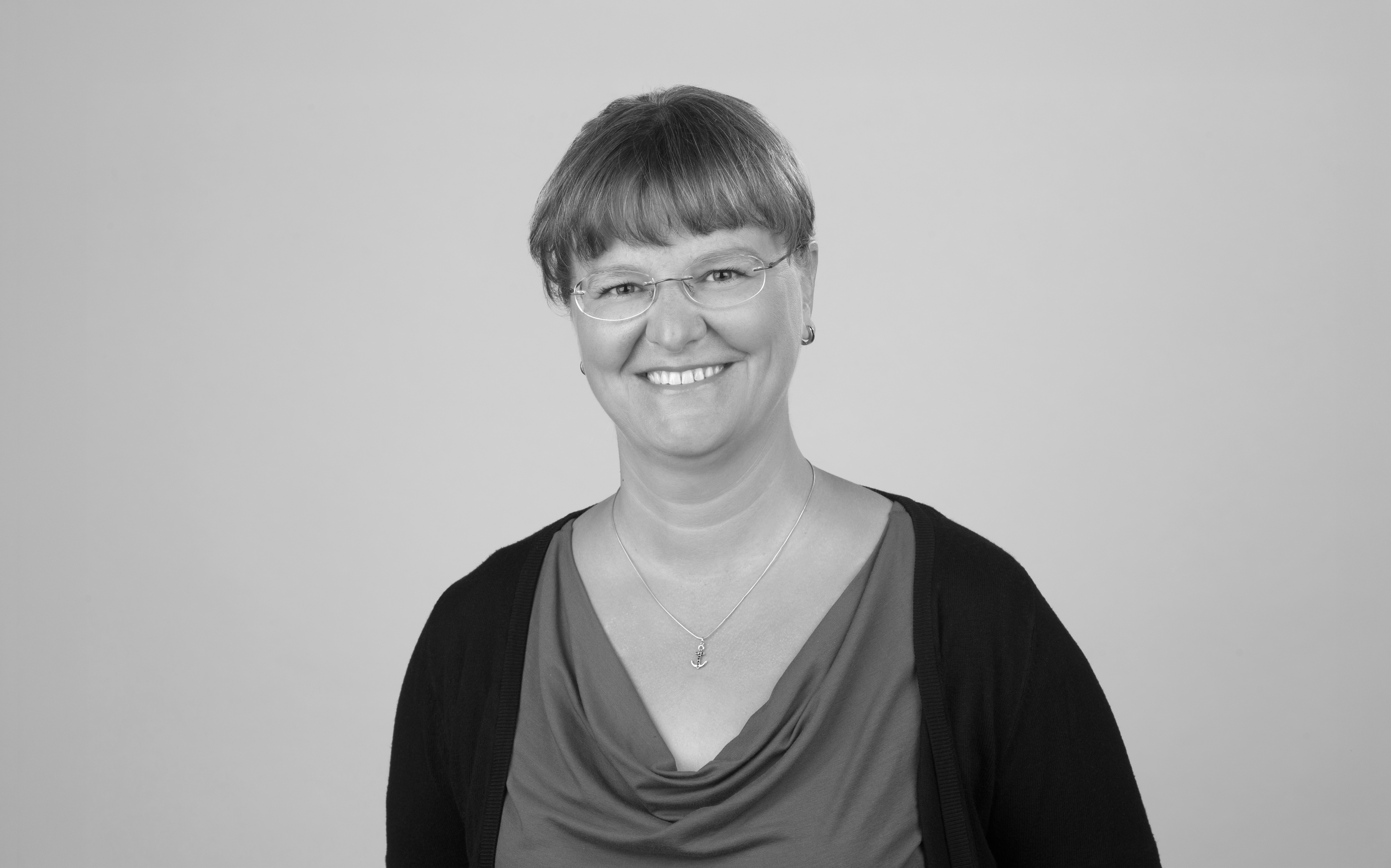 Eine lächelnde Frau mit kurzen, grauen Haaren und Brille trägt ein rotes Oberteil und einen schwarzen Cardigan. Sie steht vor einem neutralen, hellgrauen Hintergrund.