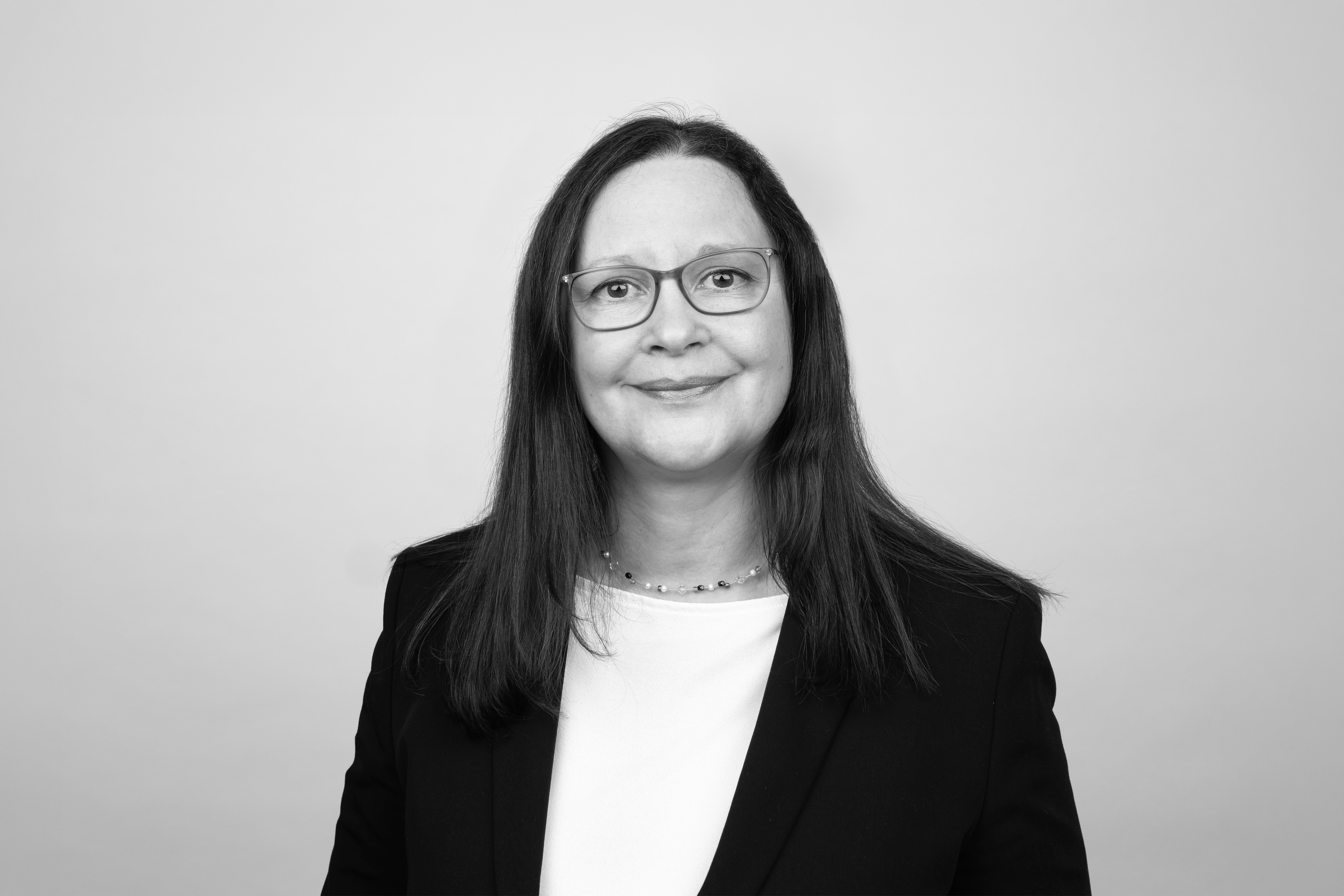 Eine Person mit langen, glatten, braunen Haaren und Brille lächelt freundlich in die Kamera, trägt einen schwarzen Blazer und ein weißes Oberteil vor einem neutralen Hintergrund.