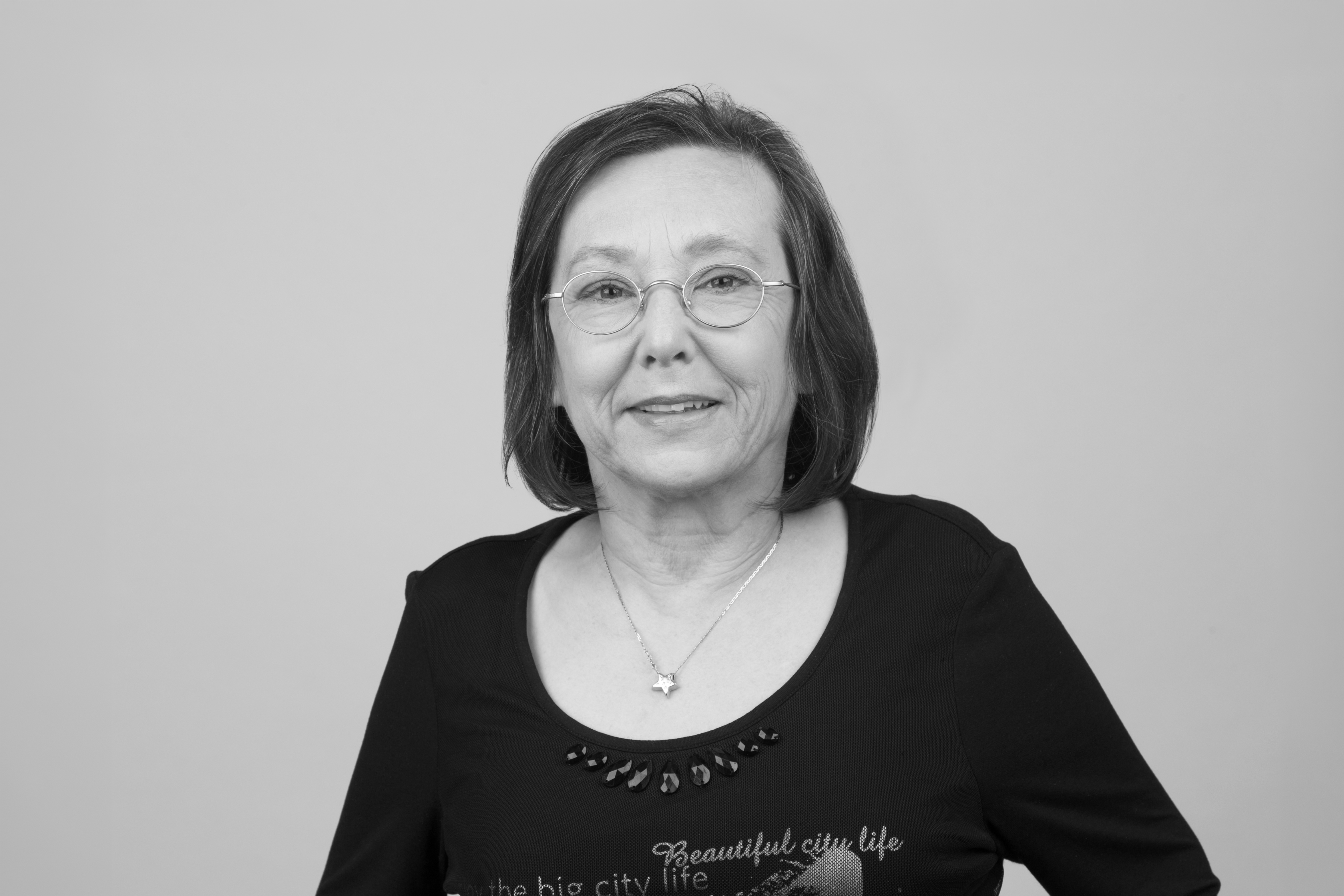 Eine lächelnde ältere Frau mit kurzen, grauen Haaren trägt eine schwarze Bluse mit Schriftzug und Schmuck. Der Hintergrund ist neutral grau.