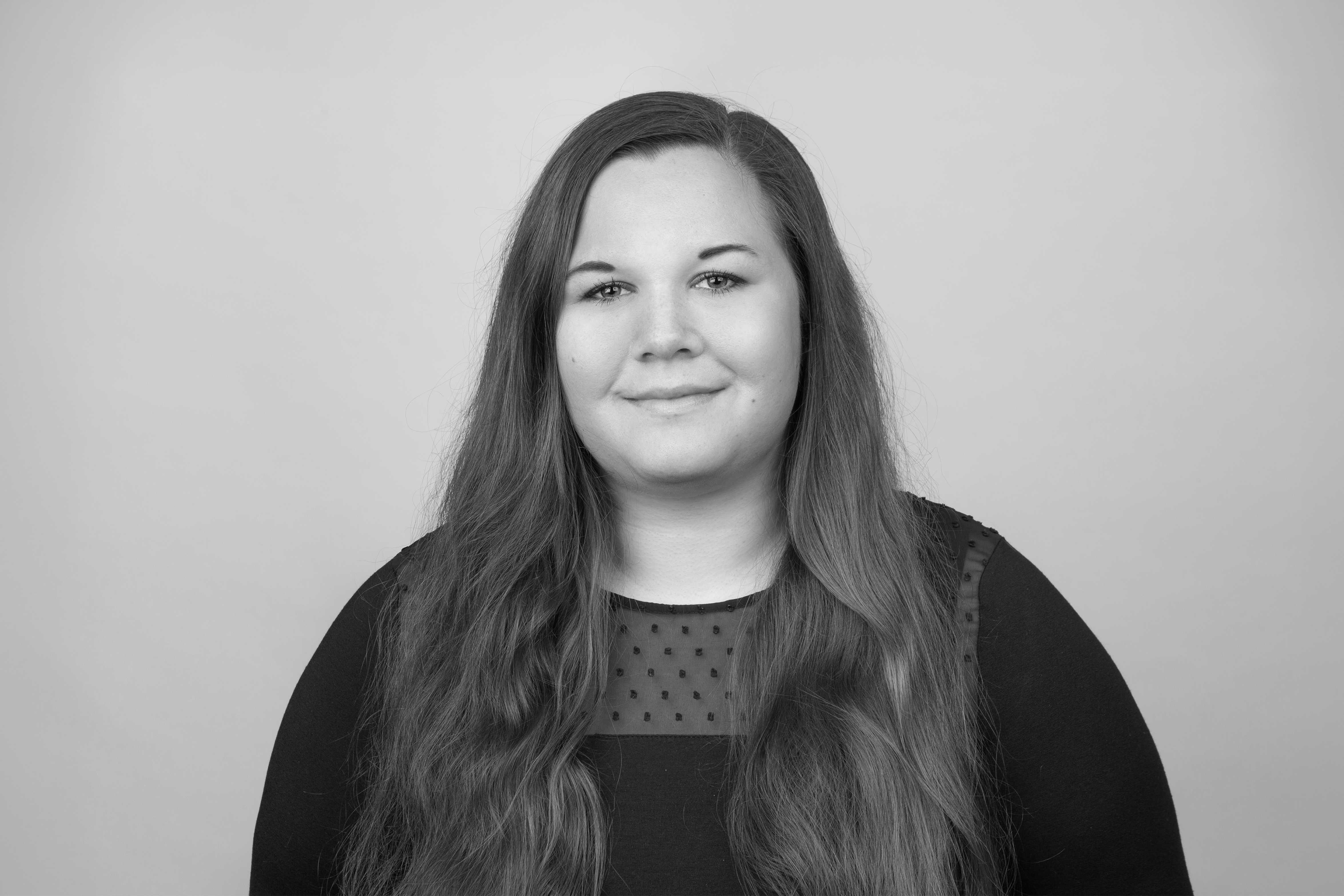 Eine lächelnde Frau mit langen, braunen Haaren trägt ein dunkelblaues Oberteil mit transparenten Ärmeln. Der Hintergrund ist hellgrau.