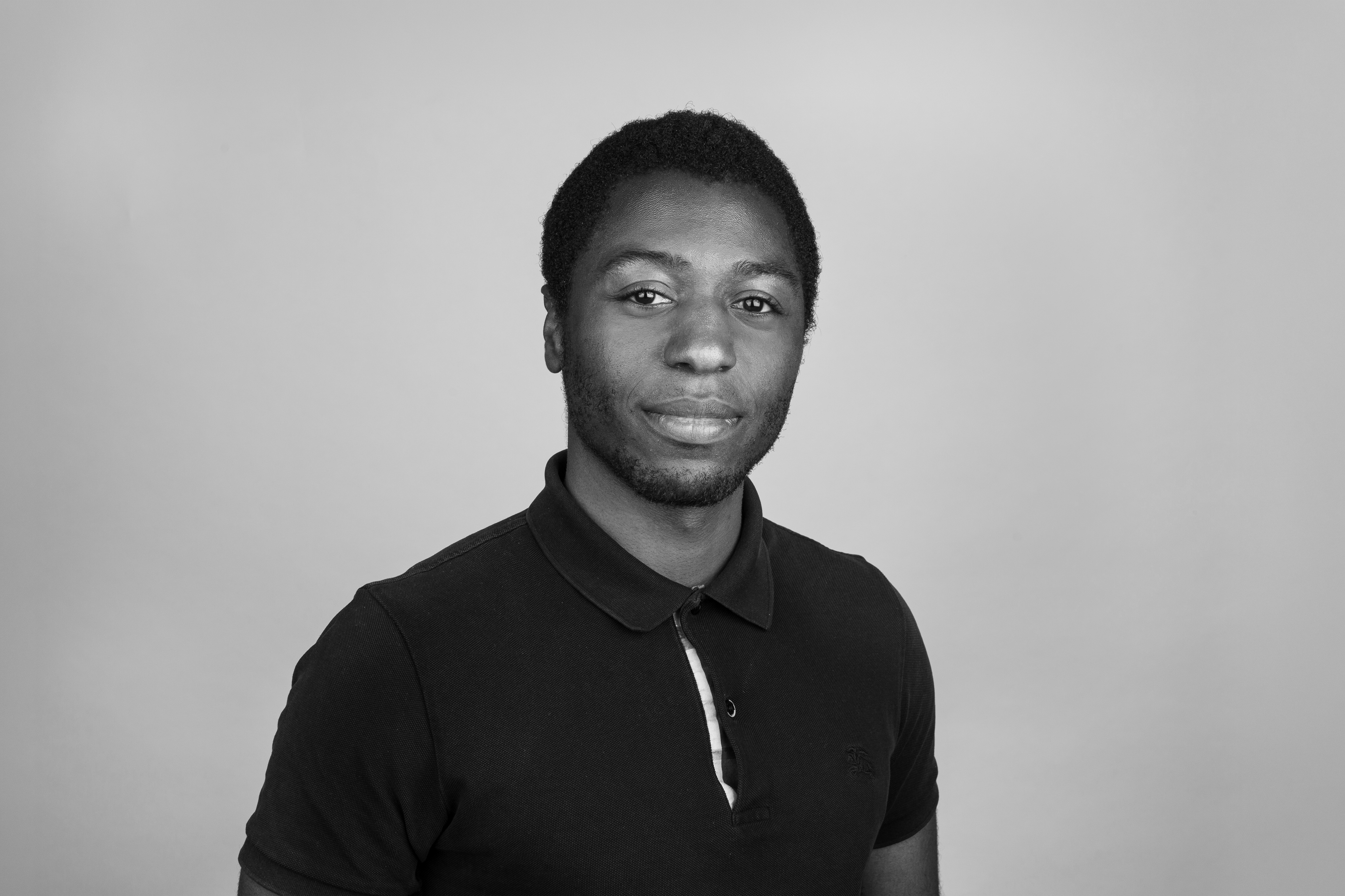Ein junger Mann mit dunklem, lockigem Haar trägt ein schwarzes Polohemd und schaut freundlich in die Kamera vor einem grauen Hintergrund.
