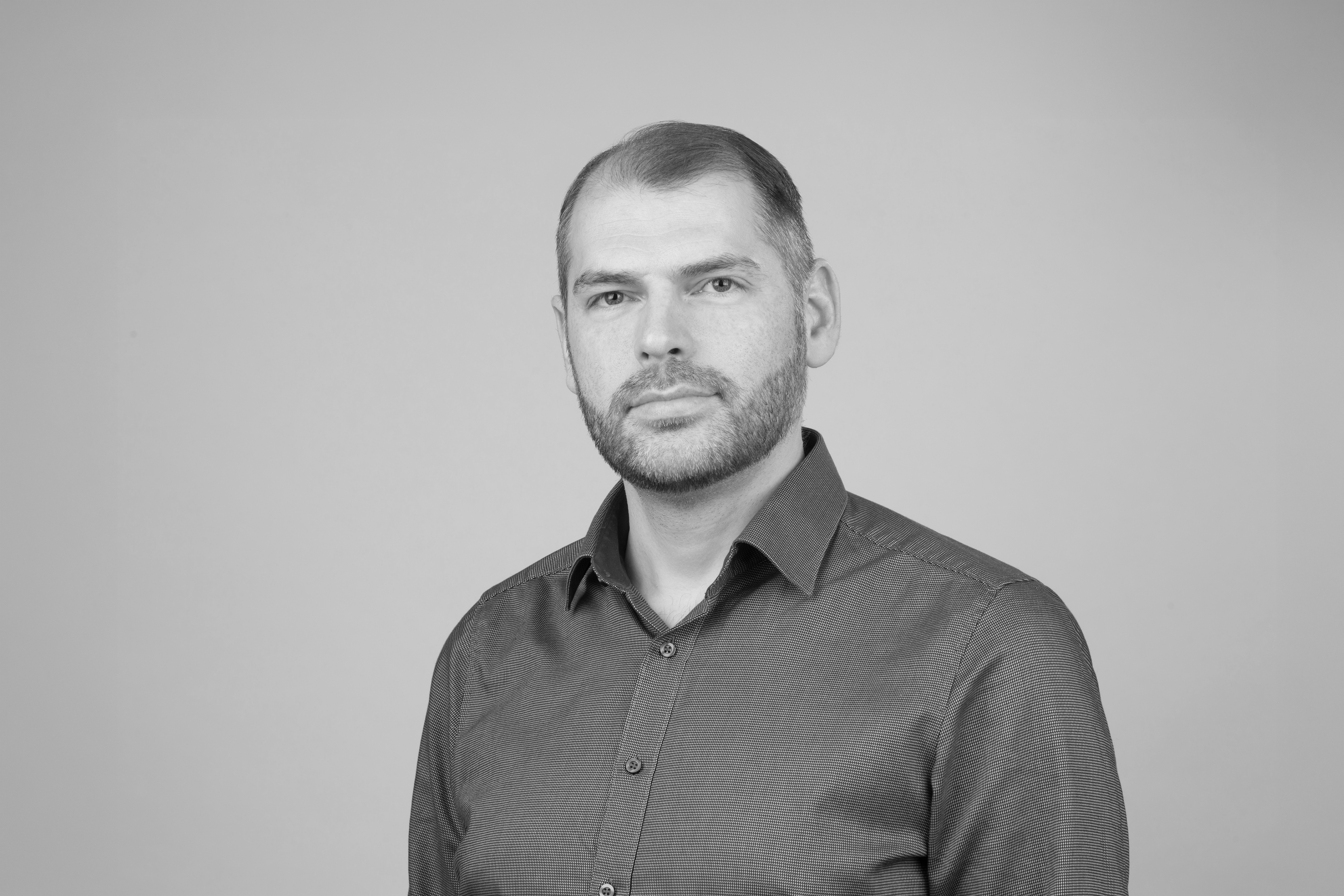 Porträt eines Mannes mit kurzem Haar, Bart und blauem Hemd vor grauem Hintergrund.