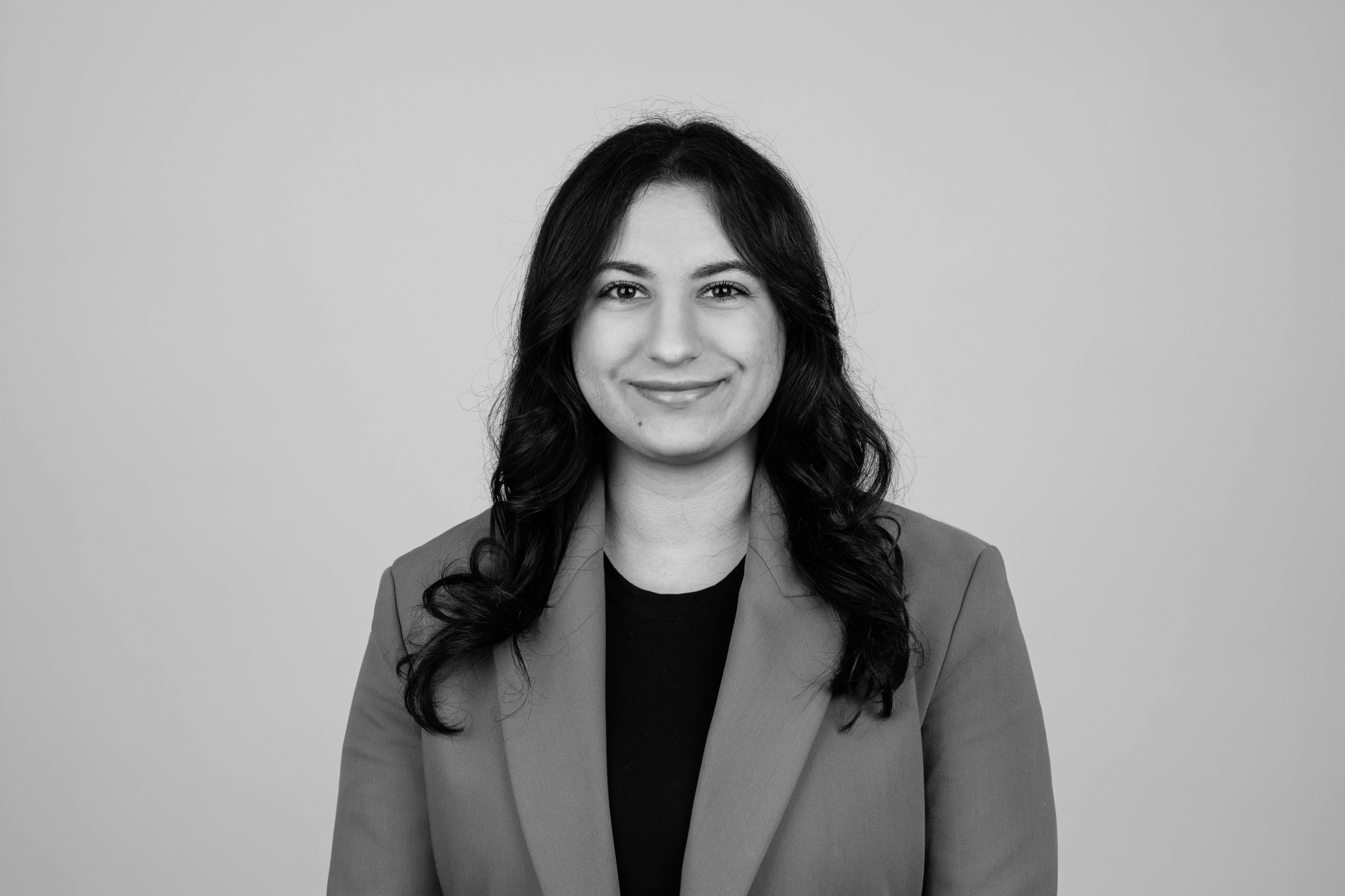 Eine lächelnde Frau mit langen, lockigen Haaren steht vor einem einfarbigen Hintergrund und trägt einen Blazer.