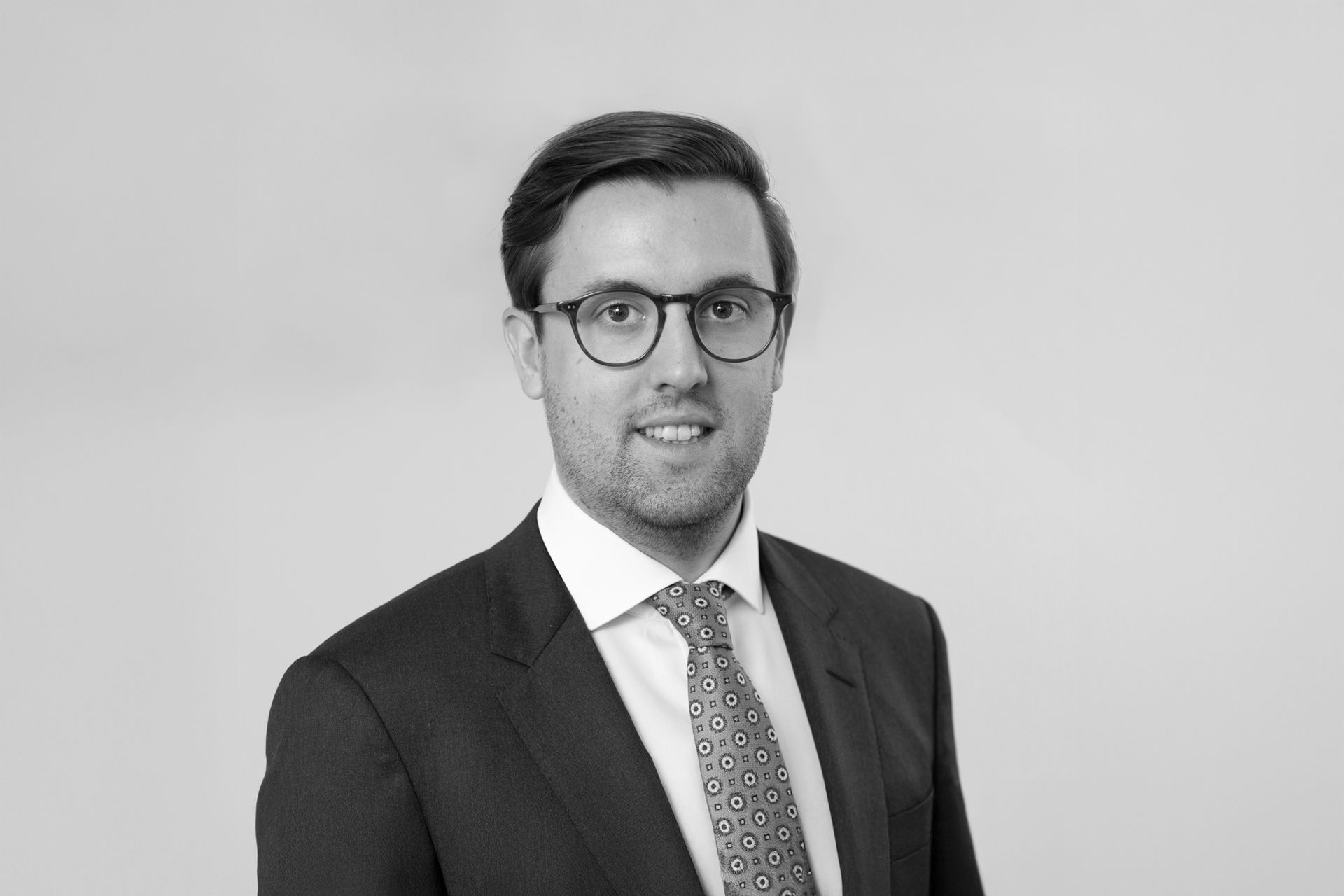 Ein gut gekleideter Mann mit Brille trägt einen Anzug und eine Krawatte mit geometrischem Muster. Er lächelt freundlich in die Kamera, vor einem neutralen, hellen Hintergrund.