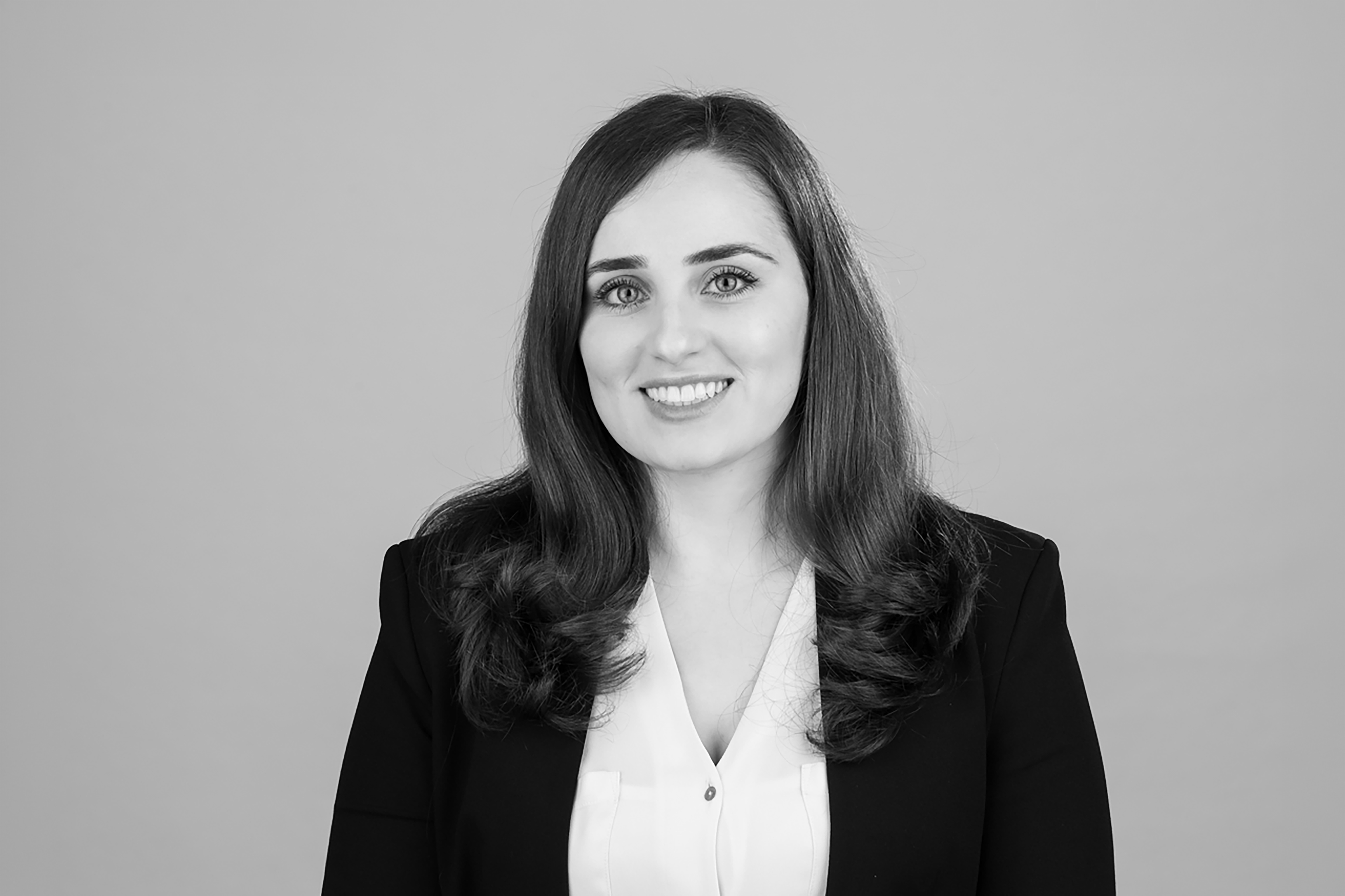 Eine lächelnde Frau mit langen, lockigen Haaren trägt ein schwarzes Blazer und ein weißes Hemd vor einem neutralen, hellen Hintergrund.