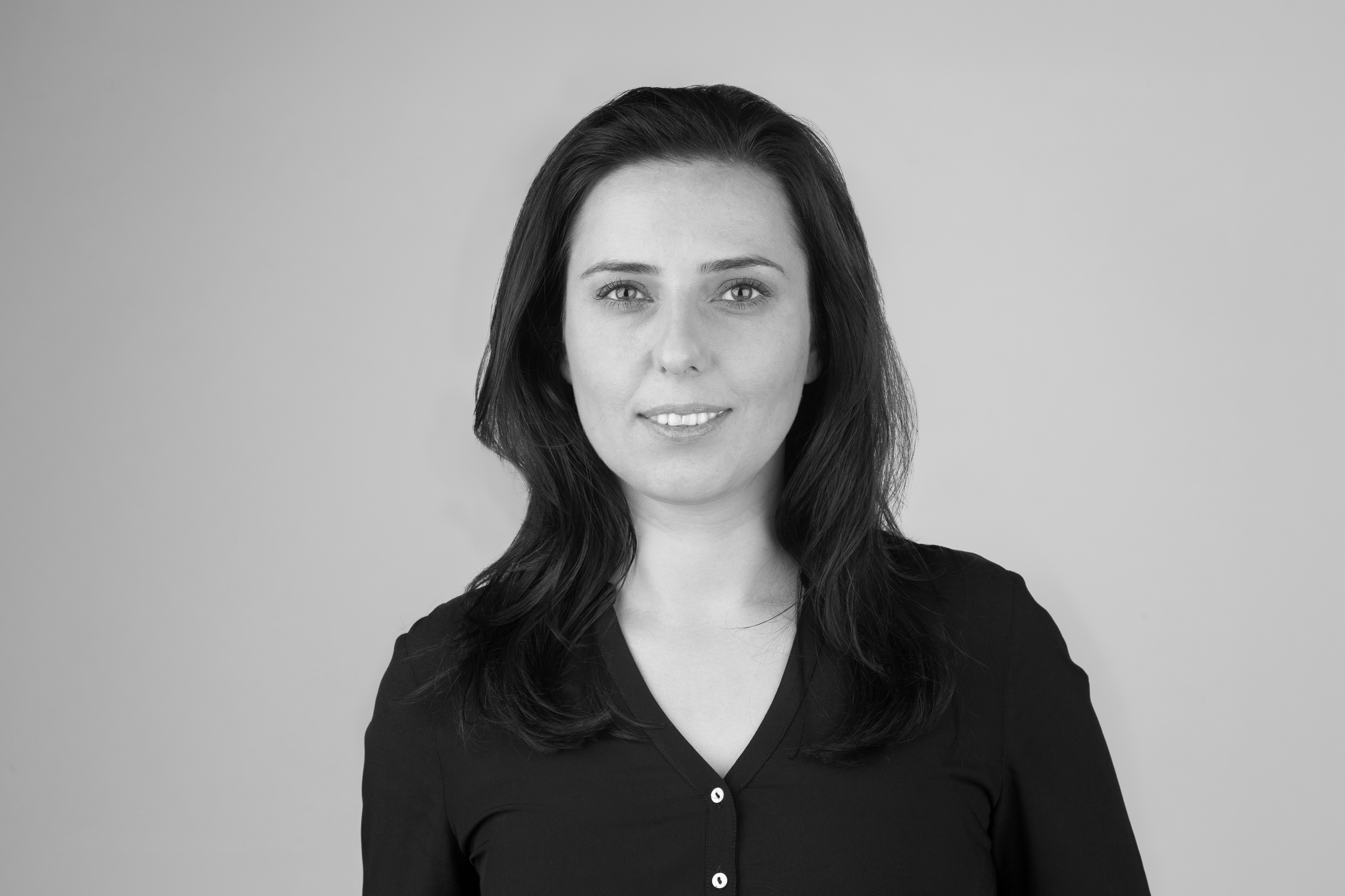 Eine Frau mit langen, dunklen Haaren und einem freundlichen Lächeln trägt ein schwarzes Oberteil. Der Hintergrund ist neutral und grau.