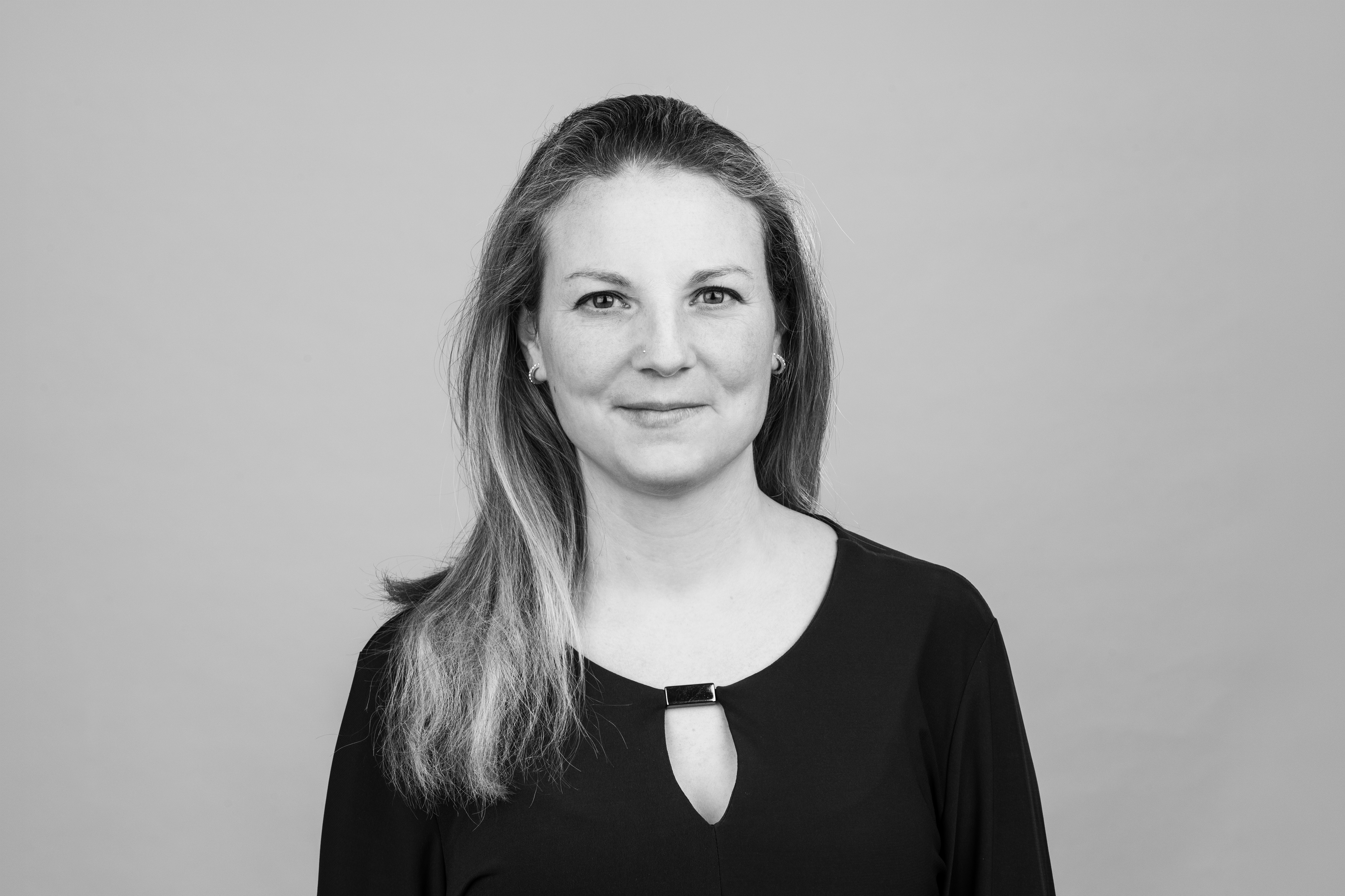 Eine Frau mit langen, hellen Haaren steht vor einem einfarbigen Hintergrund und schaut direkt in die Kamera. Sie trägt ein schwarzes Oberteil mit einer kleinen Öffnung in der Mitte.