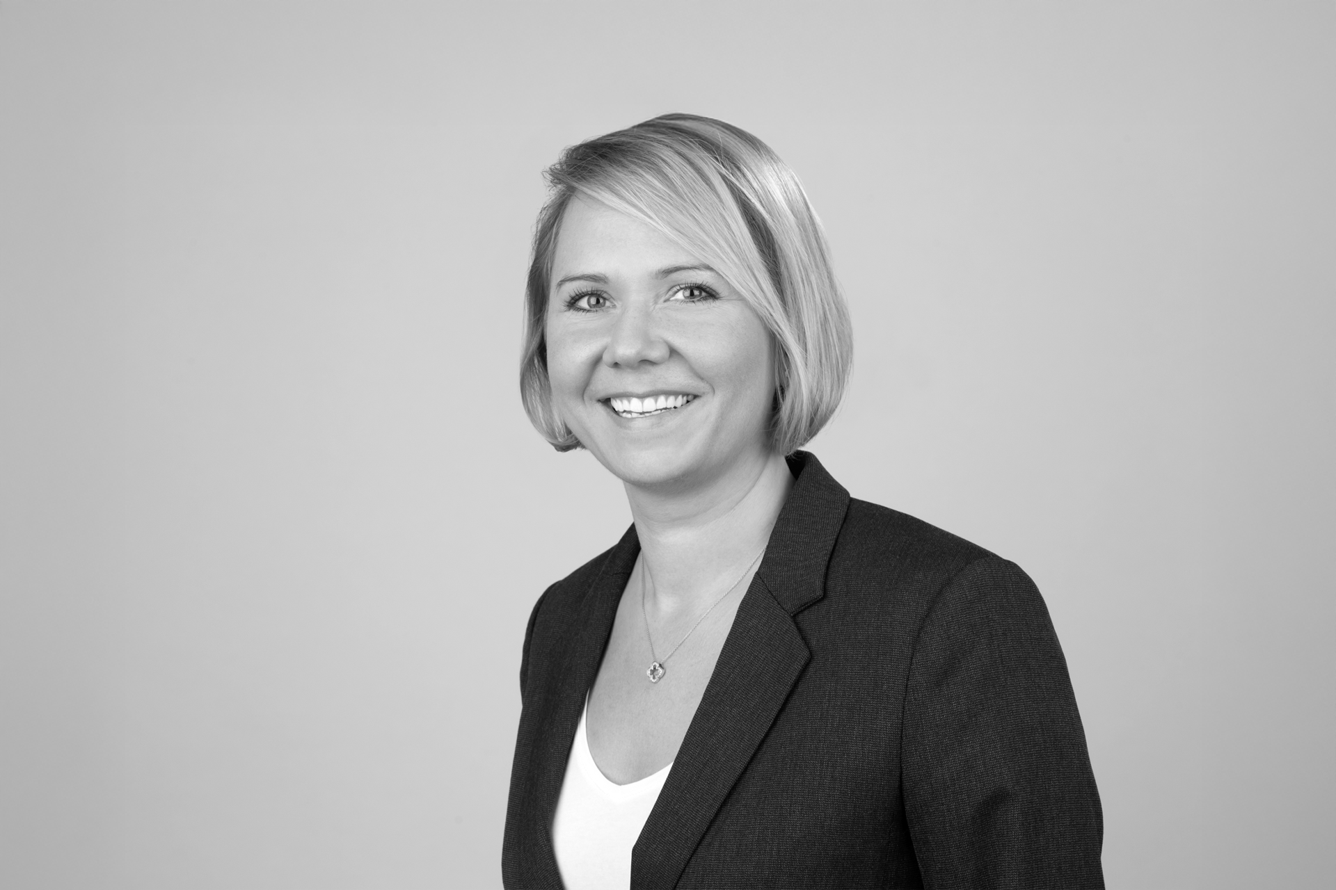 Eine lächelnde Frau mit blonden, schulterlangen Haaren trägt ein schwarzes Blazer-Jackett und eine weiße Bluse. Der Hintergrund ist grau.