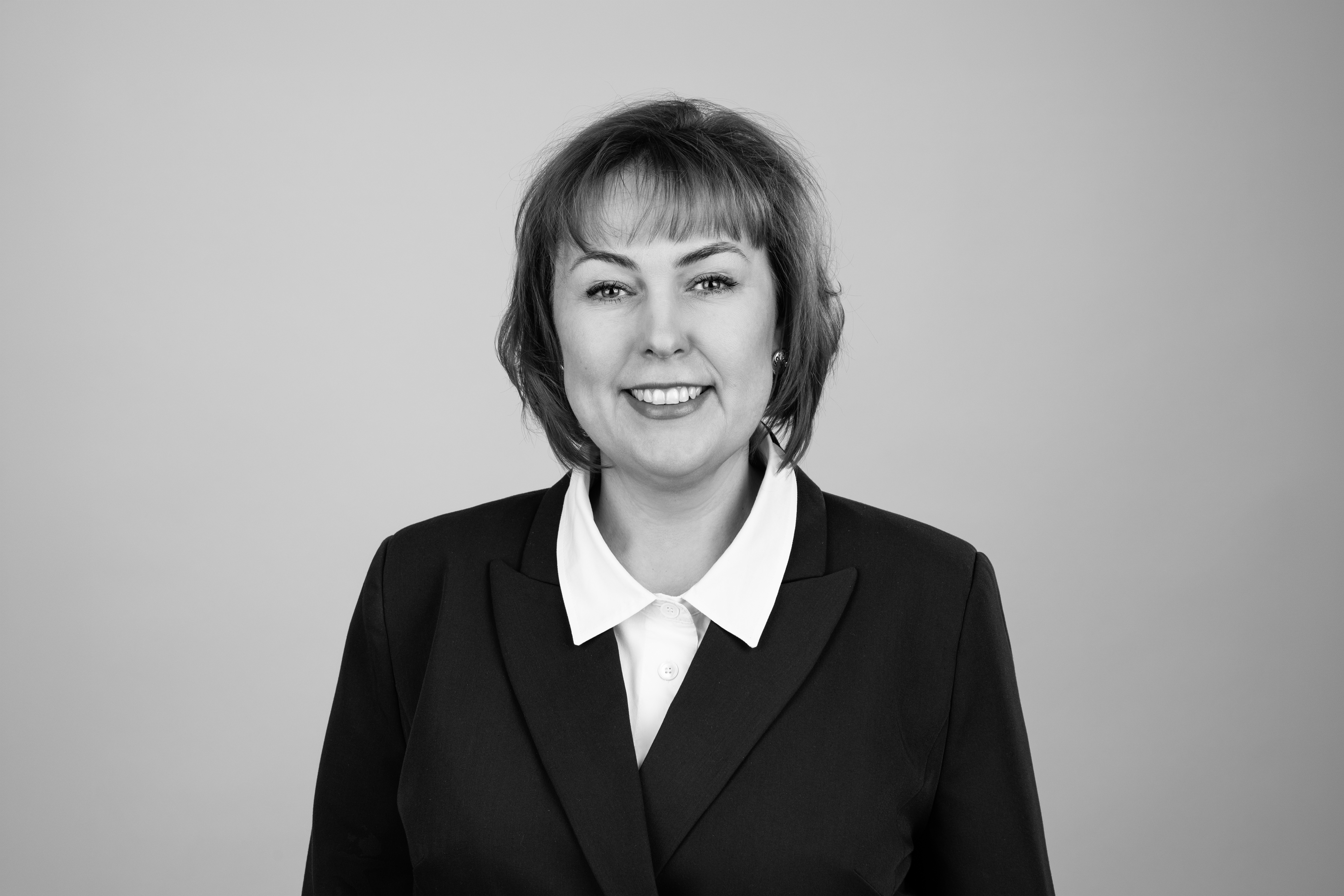 Eine lächelnde Frau mit schulterlangen, braunen Haaren trägt einen schwarzen Blazer und ein weißes Hemd. Der Hintergrund ist einfarbig und hell.