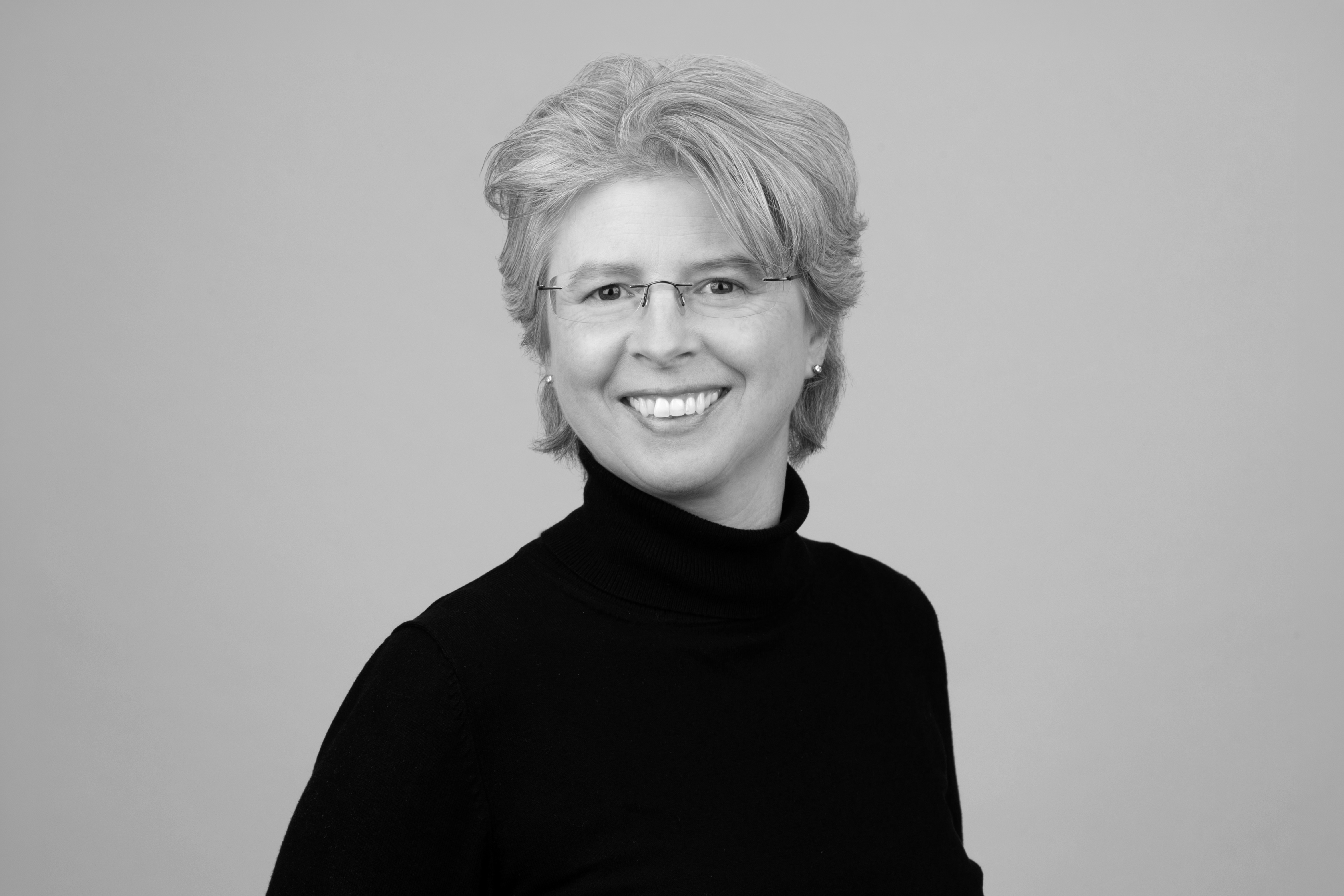 Eine lächelnde Frau mit kurzem, blonden Haar und Brille trägt einen schwarzen Rollkragenpullover und steht vor einem grauen Hintergrund.