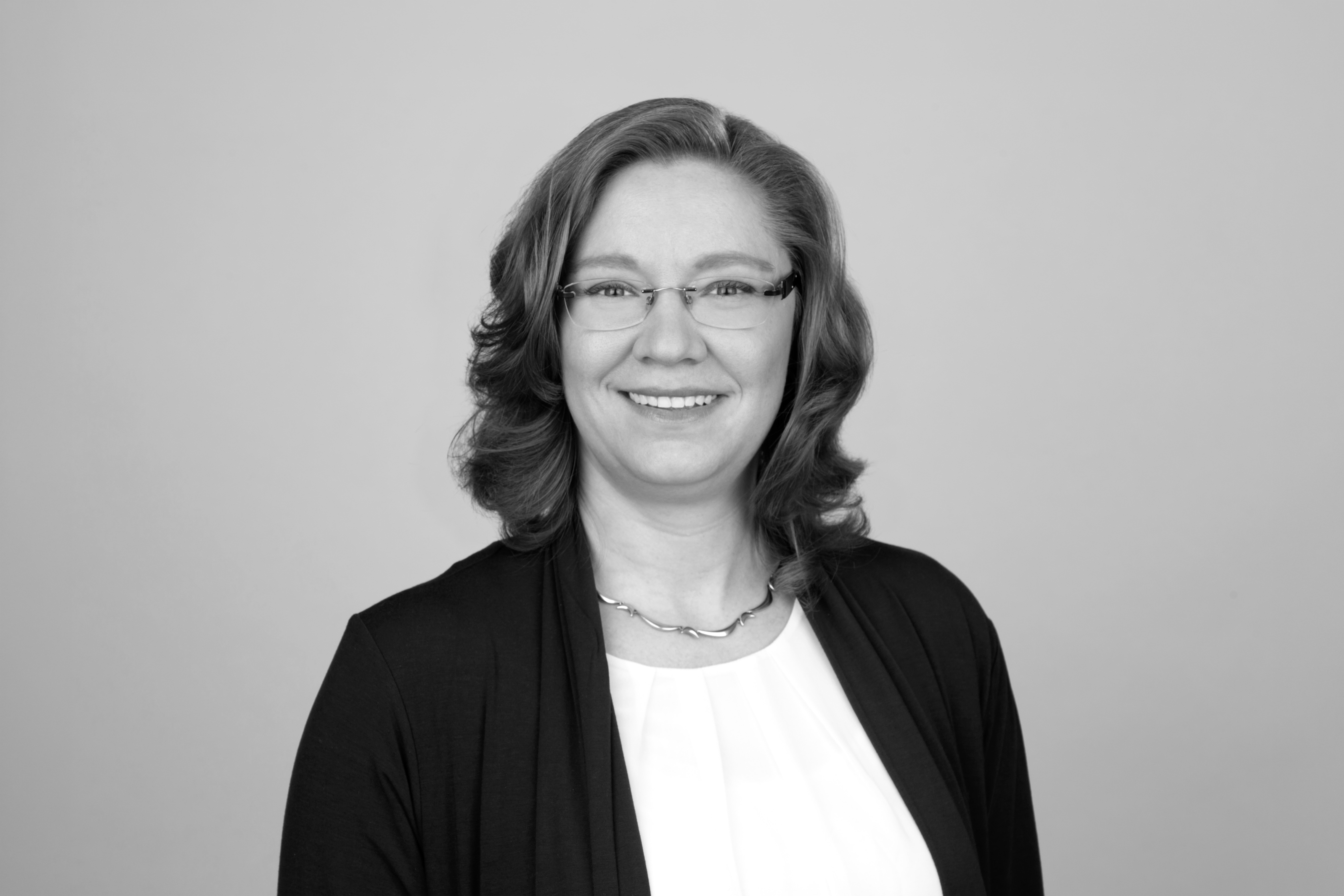 Eine freundlich lächelnde Frau mit langen, lockigen Haaren trägt eine weiße Bluse und einen dunklen Cardigan. Sie steht vor einem neutralen, hellgrauen Hintergrund.