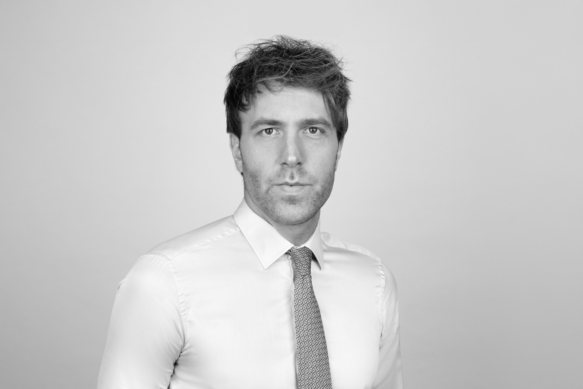 Ein junger Mann in einem hellen Hemd mit einem gestreiften roten Krawatte steht vor einem grauen Hintergrund und schaut direkt in die Kamera.