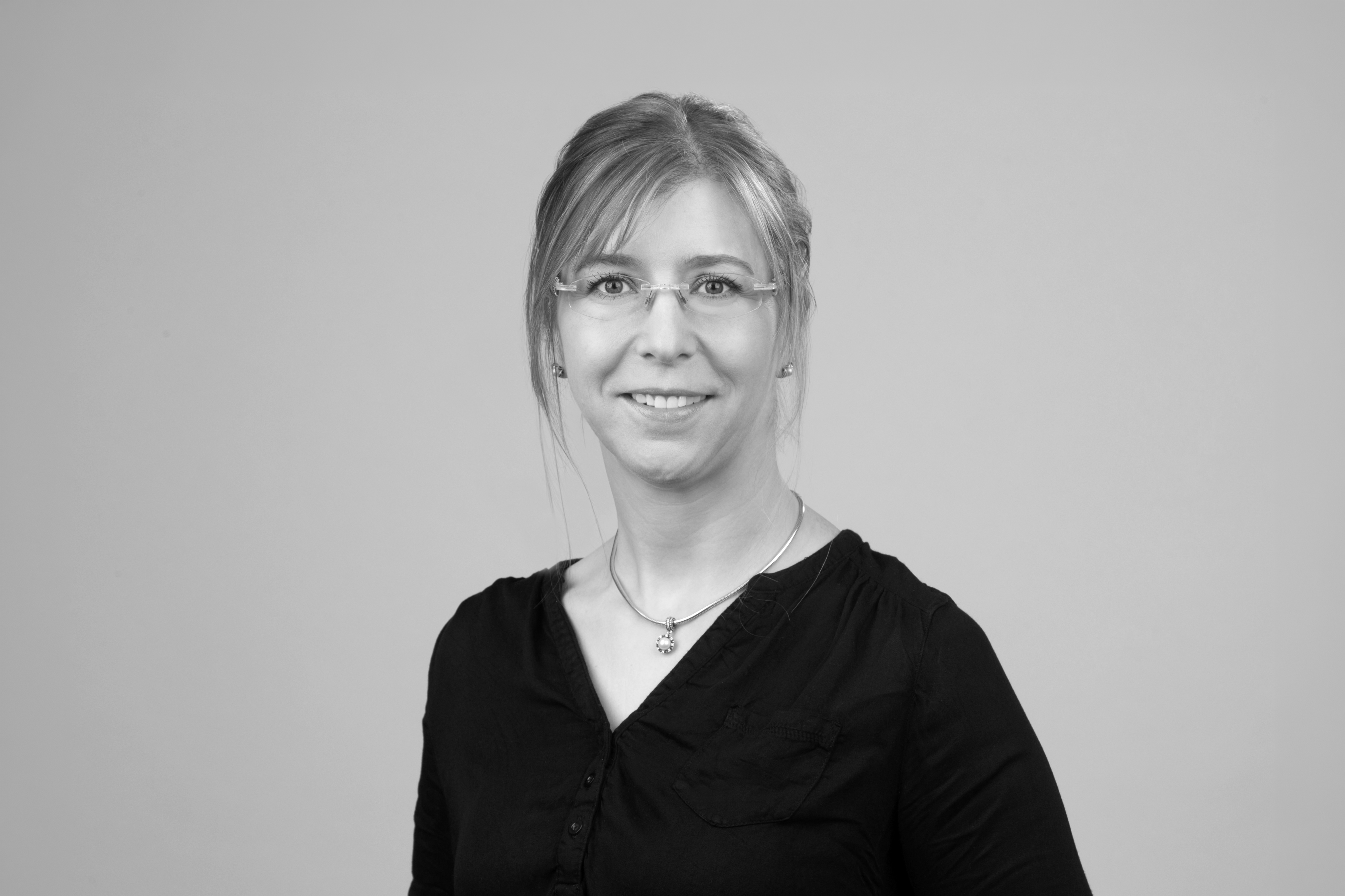 Eine Person mit langen, blonden Haaren und Brille trägt ein schwarzes Oberteil und lächelt freundlich in die Kamera vor einem grauen Hintergrund.