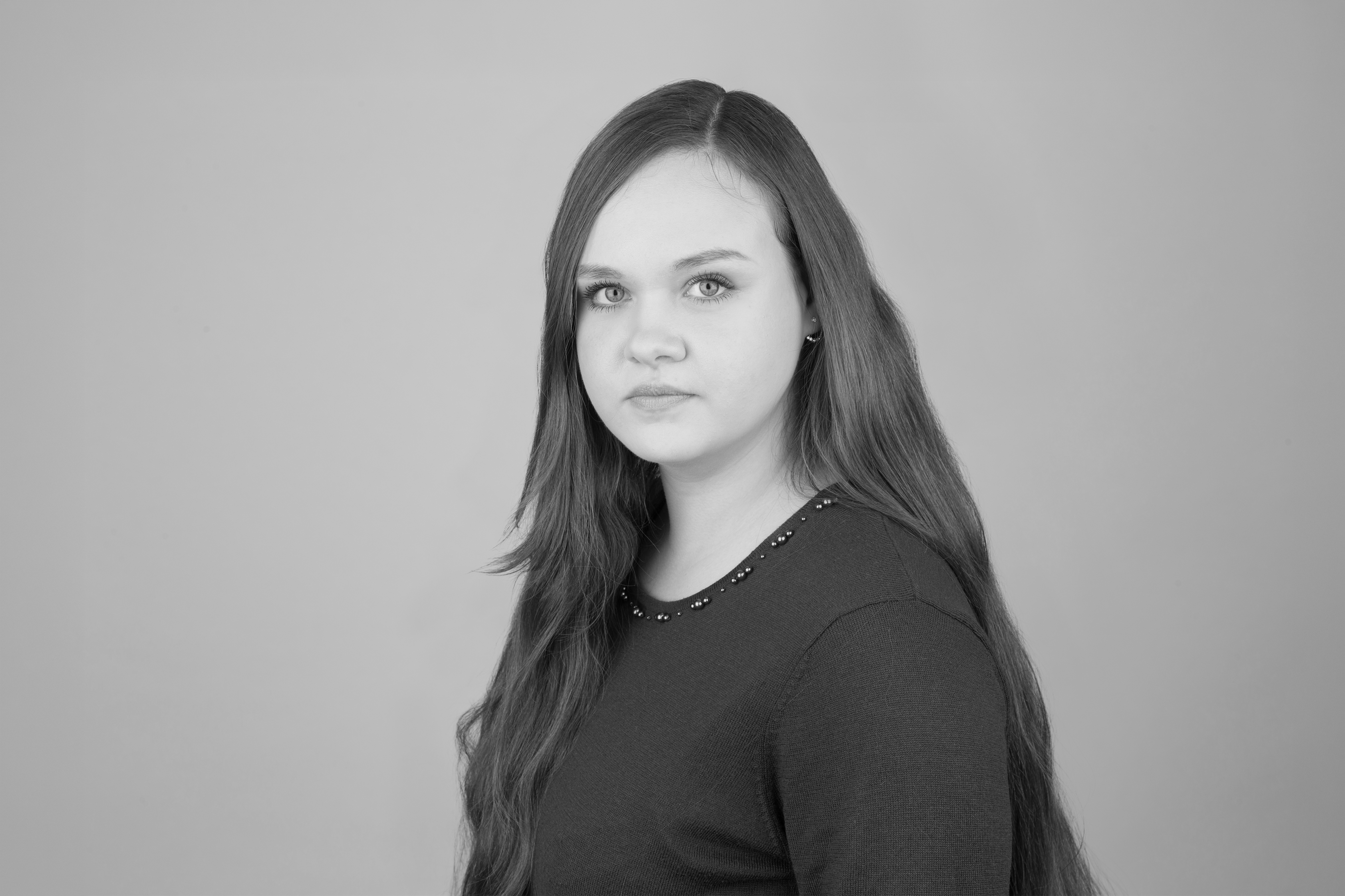 Eine junge Frau mit langen, glatten Haaren und einem neutralen Gesichtsausdruck schaut zur Seite. Sie trägt ein burgunderfarbiges Oberteil und steht vor einem hellgrauen Hintergrund.