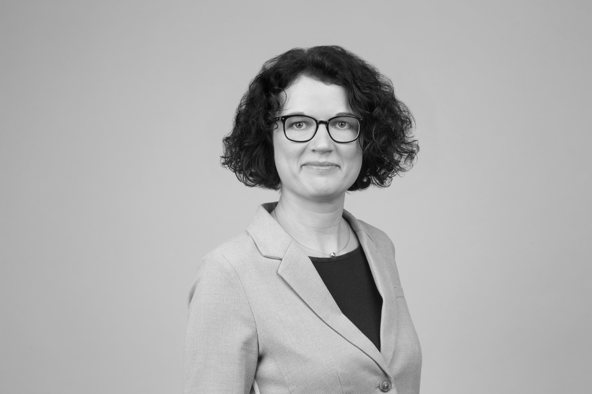 Eine Person mit lockigem, braunen Haar und Brille trägt einen grauen Blazer und blickt freundlich in die Kamera. Der Hintergrund ist neutral und einfarbig.