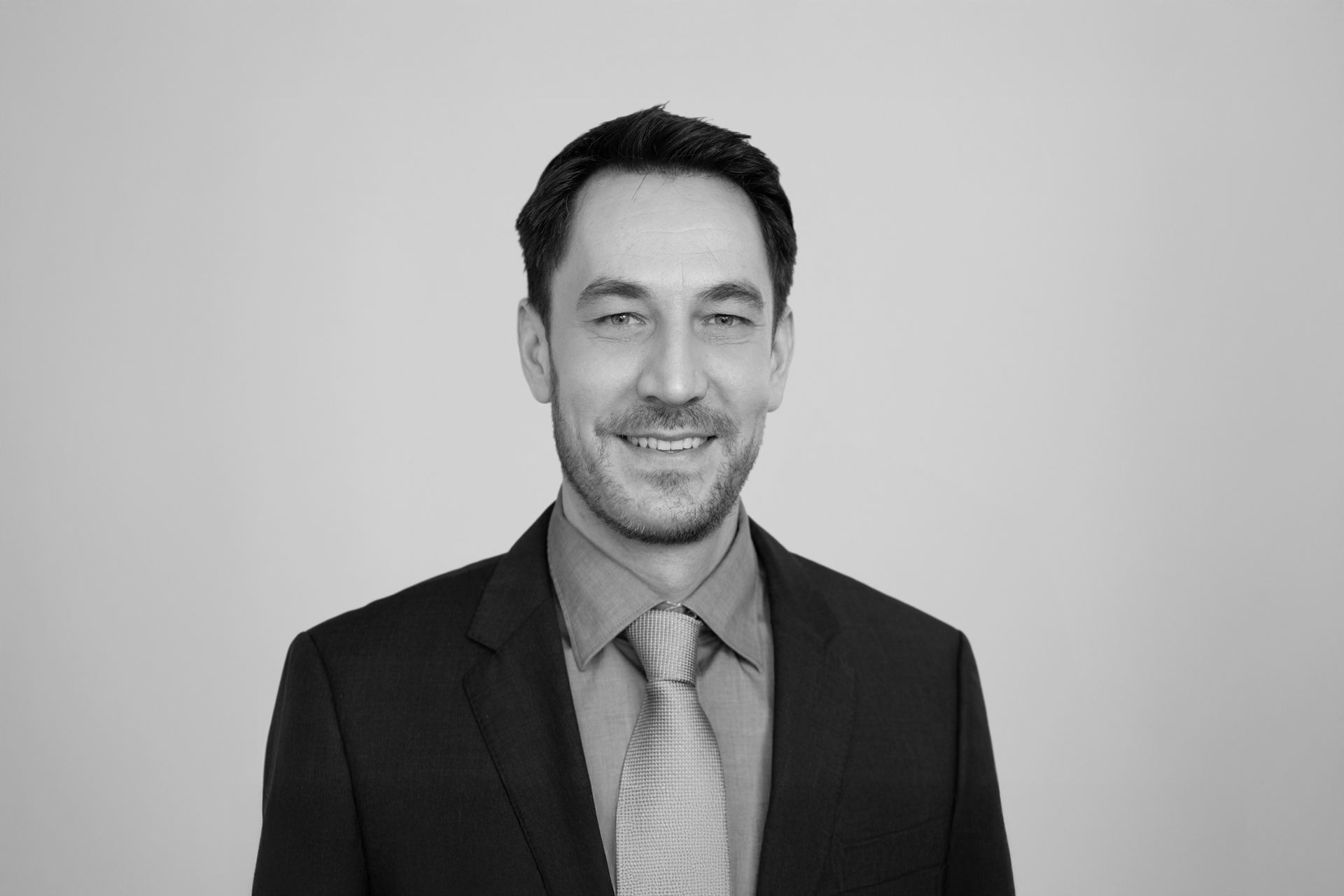 Ein Mann mit dunklen Haaren trägt einen schwarzen Anzug und ein hellblaues Hemd mit einer hellgrauen Krawatte und lächelt freundlich vor einem grauen Hintergrund.