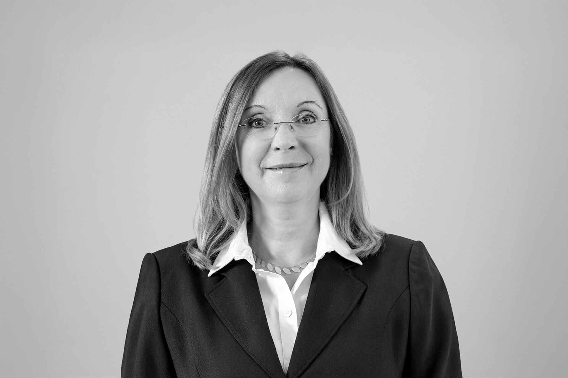 Eine Frau mit langen, blonden Haaren und Brille trägt einen schwarzen Blazer über einem weißen Hemd. Sie lächelt und steht vor einem grauen Hintergrund.